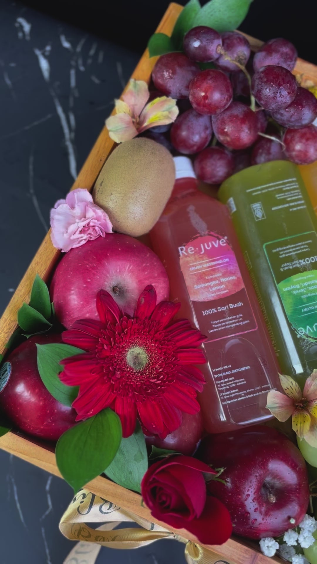 A wooden fruit box containing an assortment of fresh fruits such as apples, oranges, grapes, and bananas, along with two bottles of juice and decorative flowers. rejuve juice fruit hamper jakarta, surabaya, cirebon