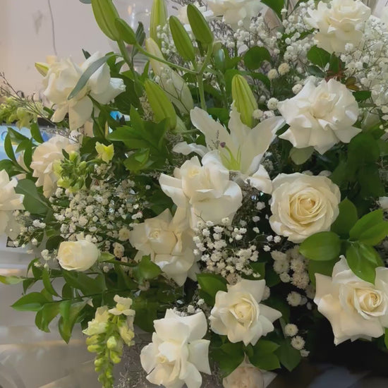 Bouquet of white flowers with greenery in a clear plastic wrap, placed indoors. Bouquet standing flower of white flowers with greenery on a stand in a store setting. florist surabaya, standing flower jakarta, standing flower surabaya