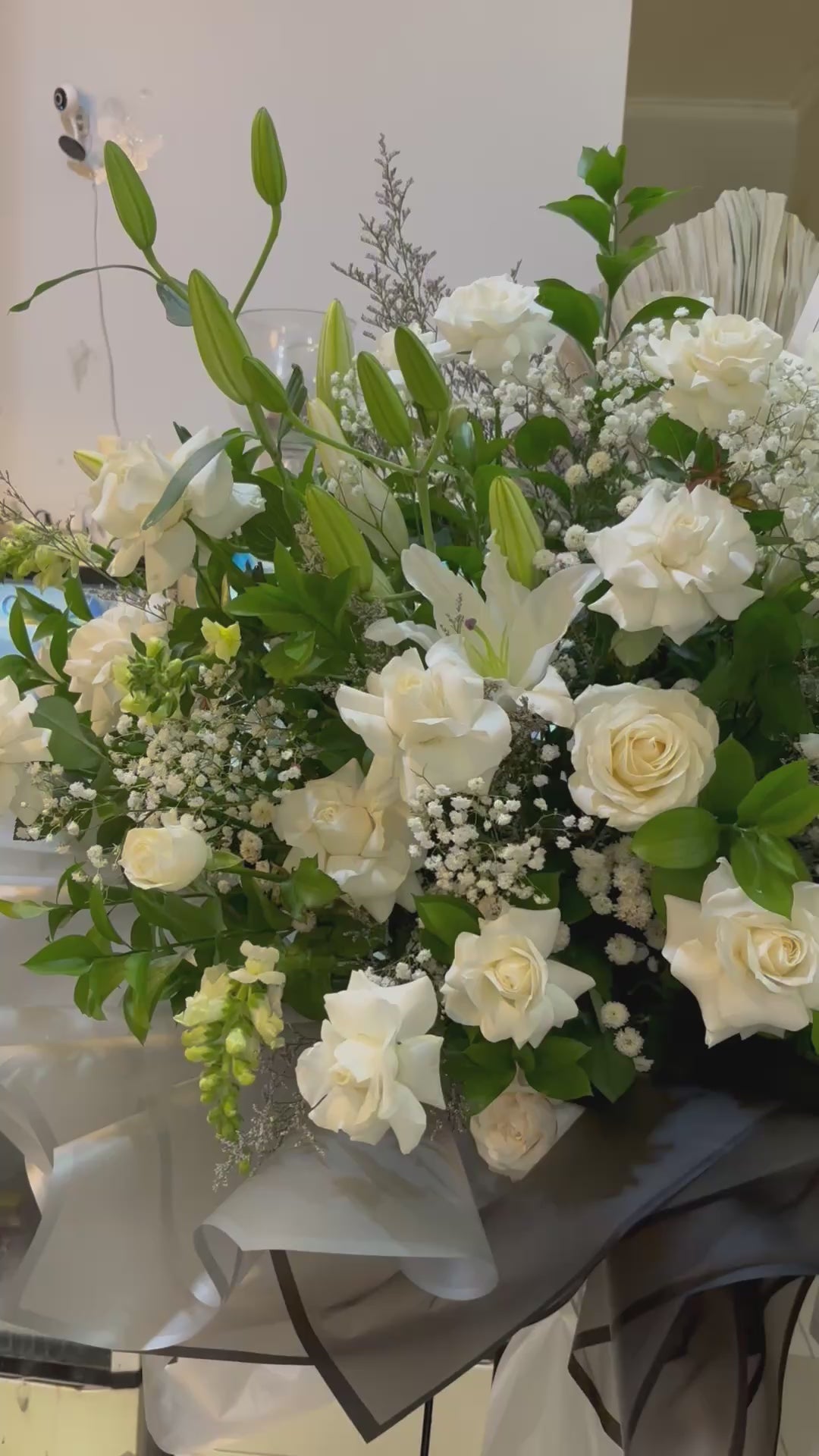 Bouquet of white flowers with greenery in a clear plastic wrap, placed indoors. Bouquet standing flower of white flowers with greenery on a stand in a store setting. florist surabaya, standing flower jakarta, standing flower surabaya